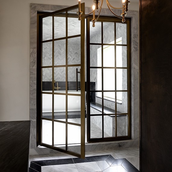 Outside view of wet room with white marble herringbone mosaic on walls, white marble rectangle mosaic, white marble hexagon mosaic on the floor, and black marble look tile surrounding the tube and on sitting bench.