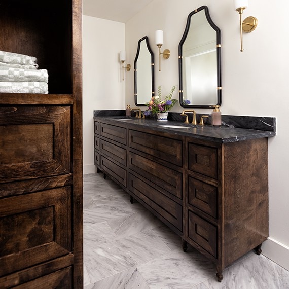 Sideview of bathroom with his and hers sinks, dark wood cabinets, gold fixtures, white marble look floor tile, and black marble countertops.