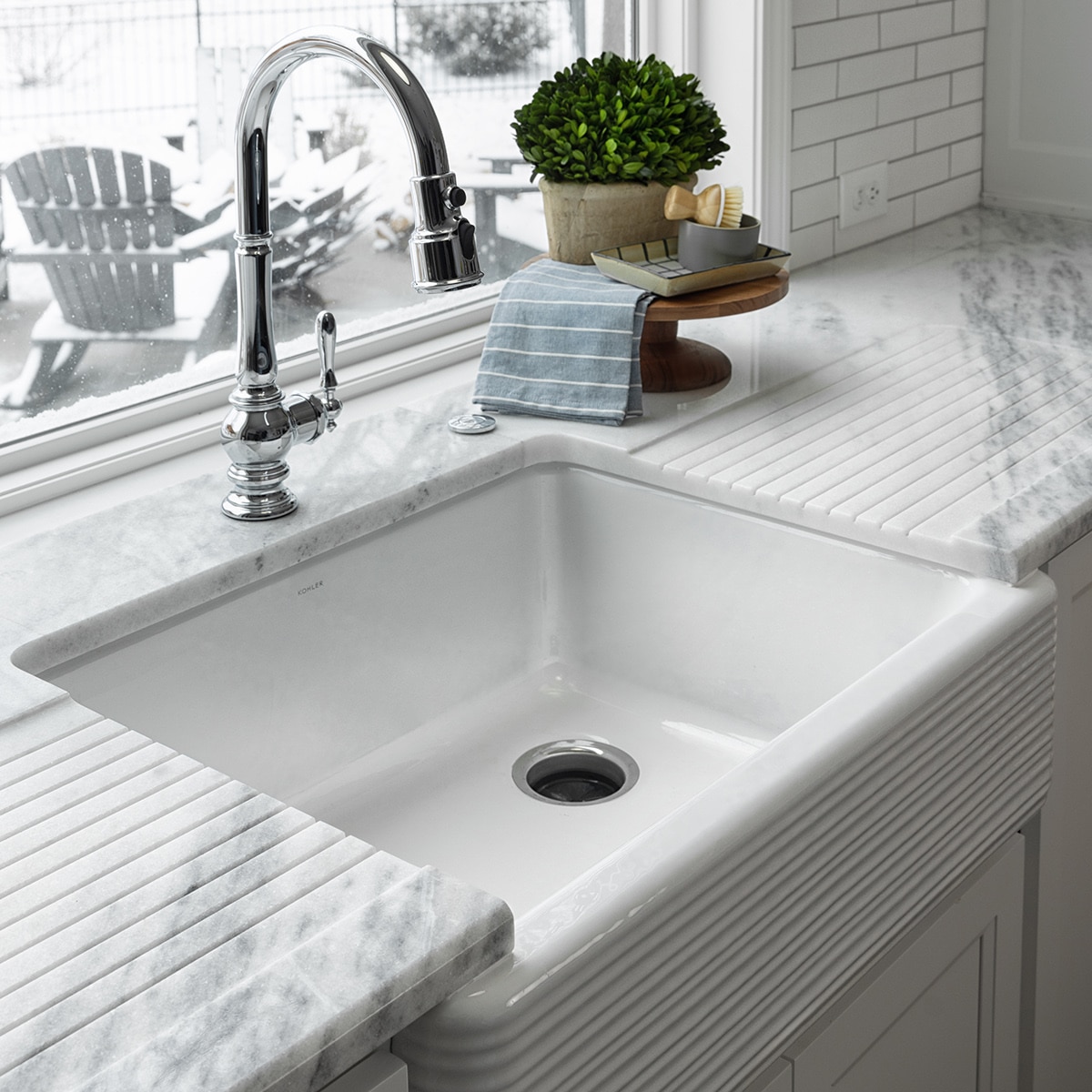 Kitchen sink with silver faucet, white cabinets, white rippled tile on the sink wall, and a white marble slab countertop.