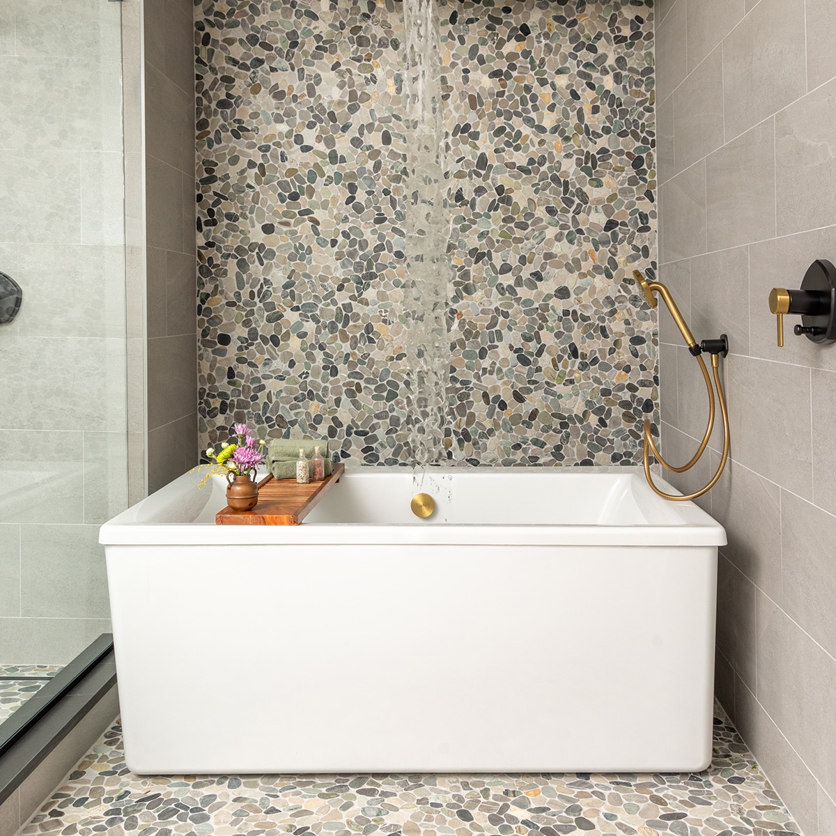 Bathtub with pebble mosaic surrounding the tub floor and back wall, and gray stone look tile on right wall.
