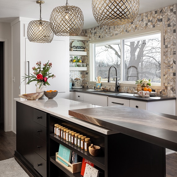 Kitchen with white cabinets, natural stone mosaic for the walls and backsplash, and an island with black cabinets and a beige marble countertop.