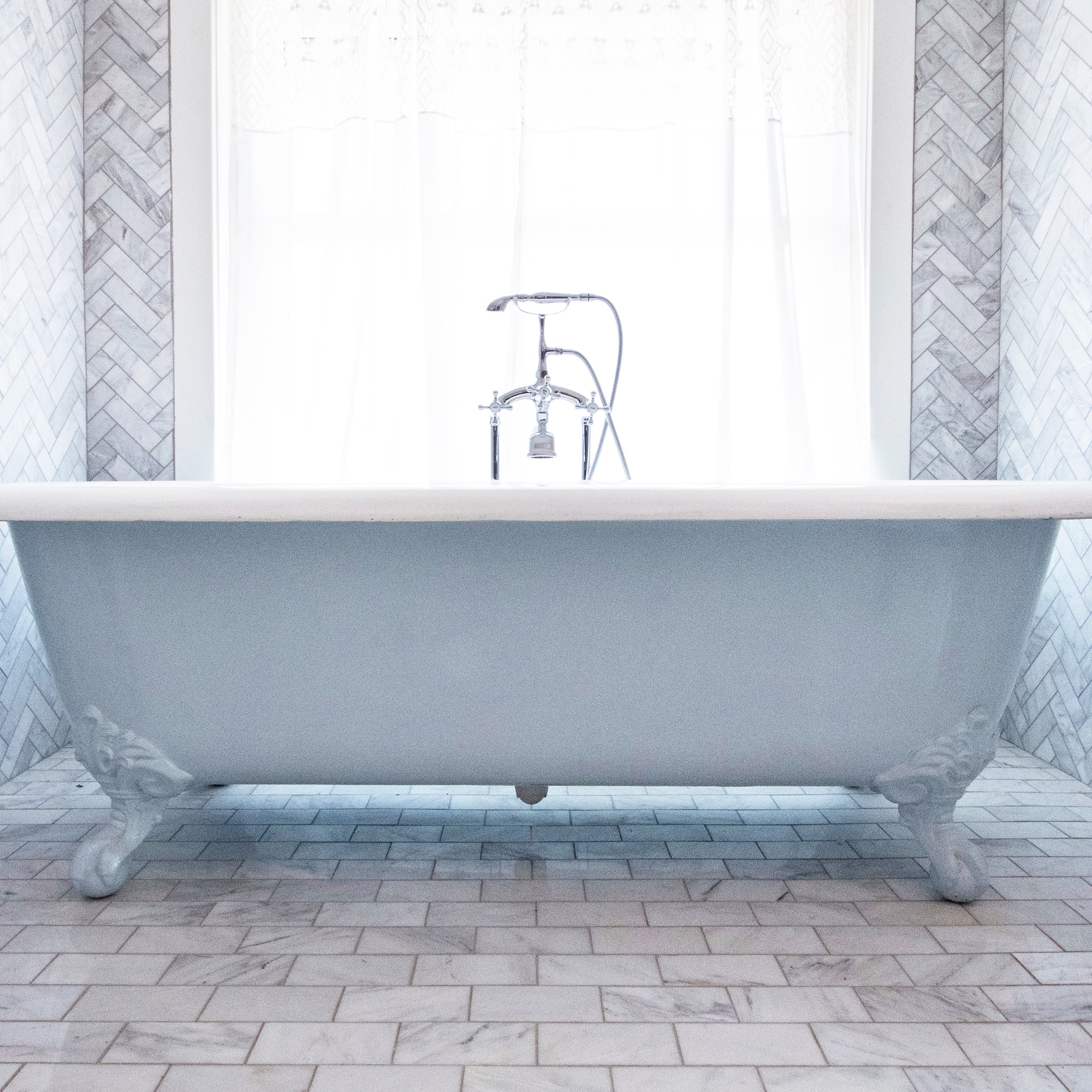 Blue bathtub with white natural stone herringbone mosaic on the walls, and white marble look brick joint tile on the floor.