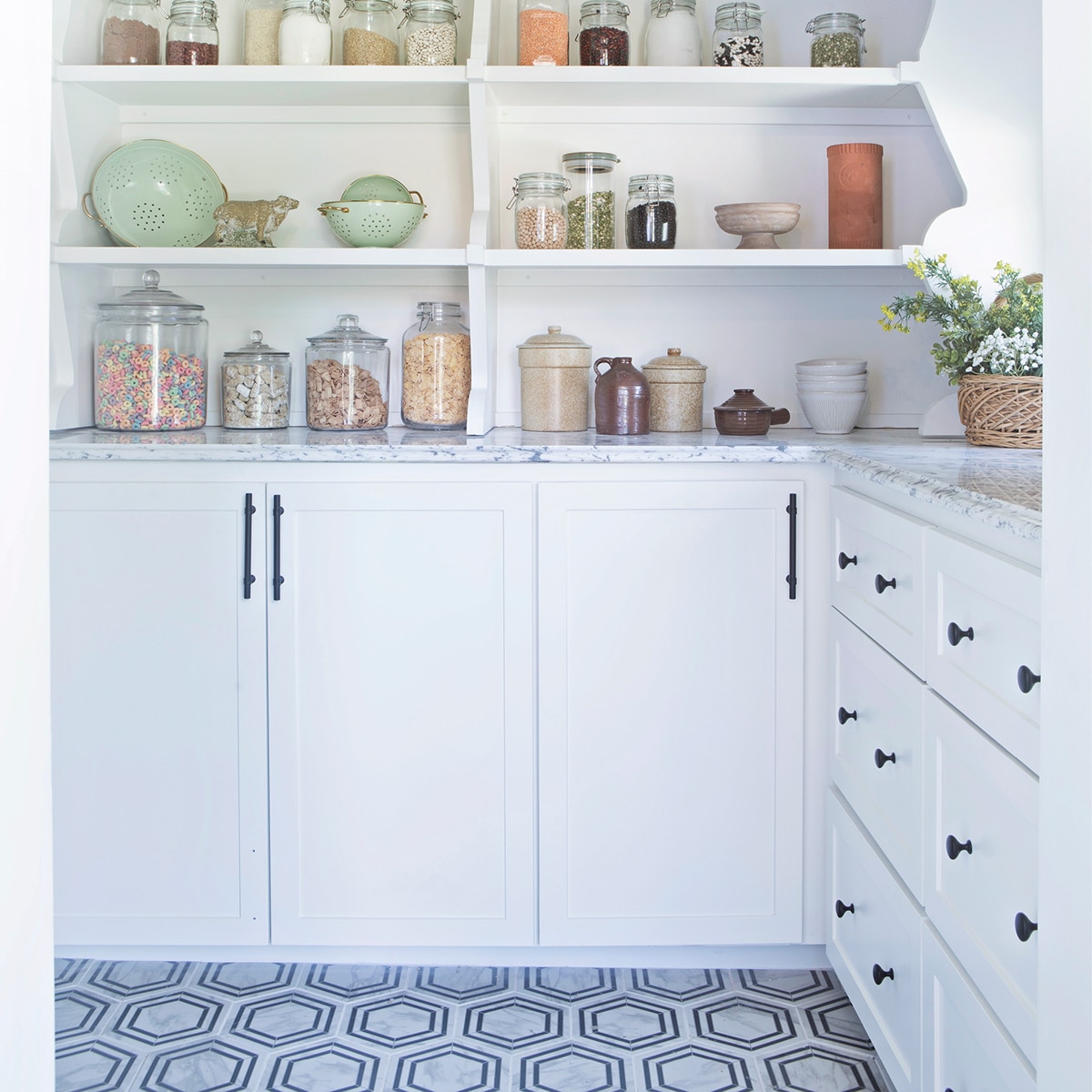 Pantry with food in jars on white shelves and drawers, white marble countertop, and  a black and white hexagon floor tile.