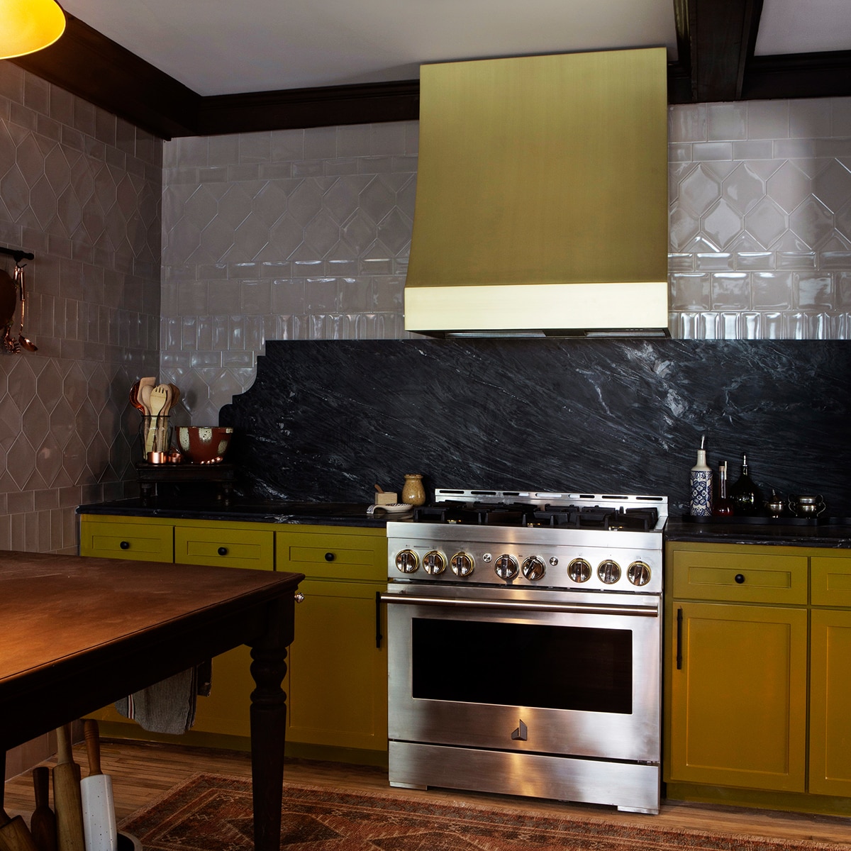 Kitchen with yellow cabinets, silver stove, light wood floors, tan tile in various sizes on the walls, and a black marble look slab countertop and backsplash.