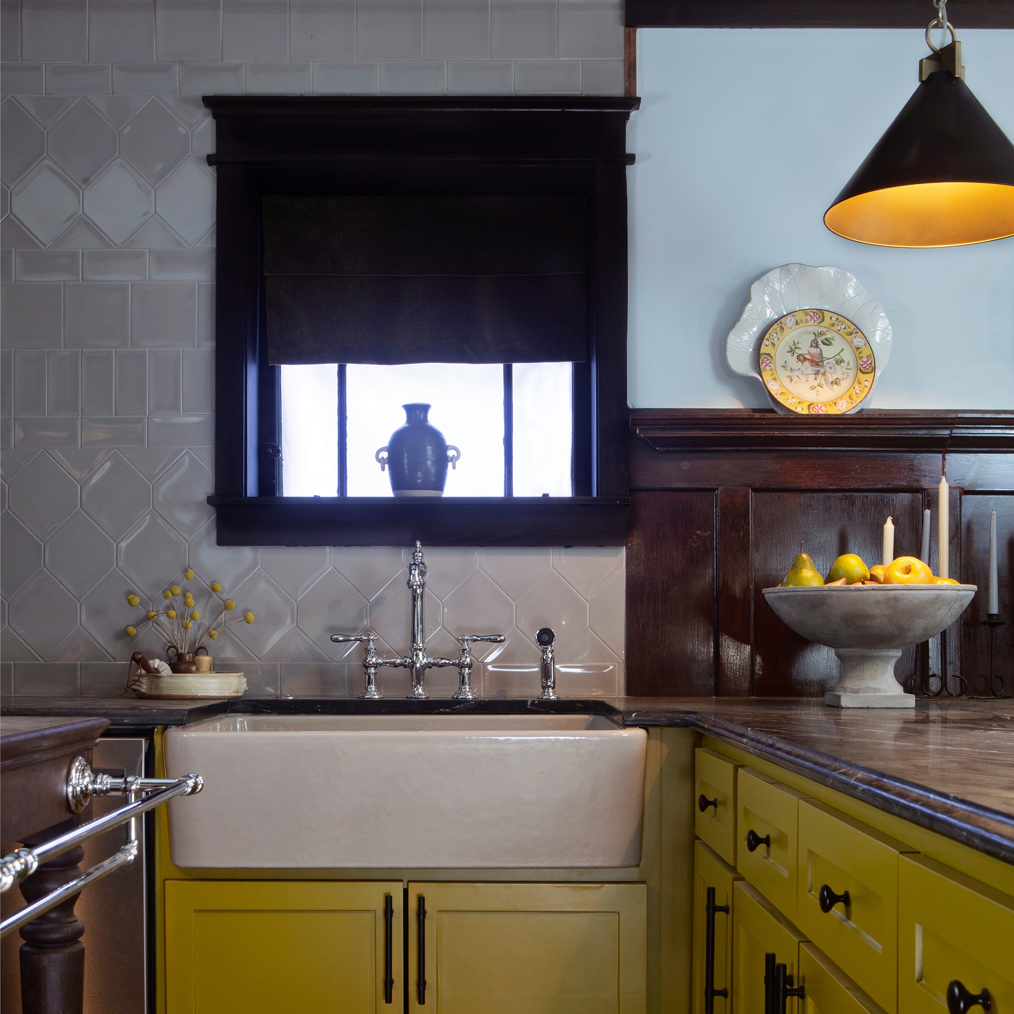 Kitchen with yellow cabinets, farmhouse style sink, light wood floors, tan tile in various sizes on the walls, and a black marble look slab countertop and backsplash.