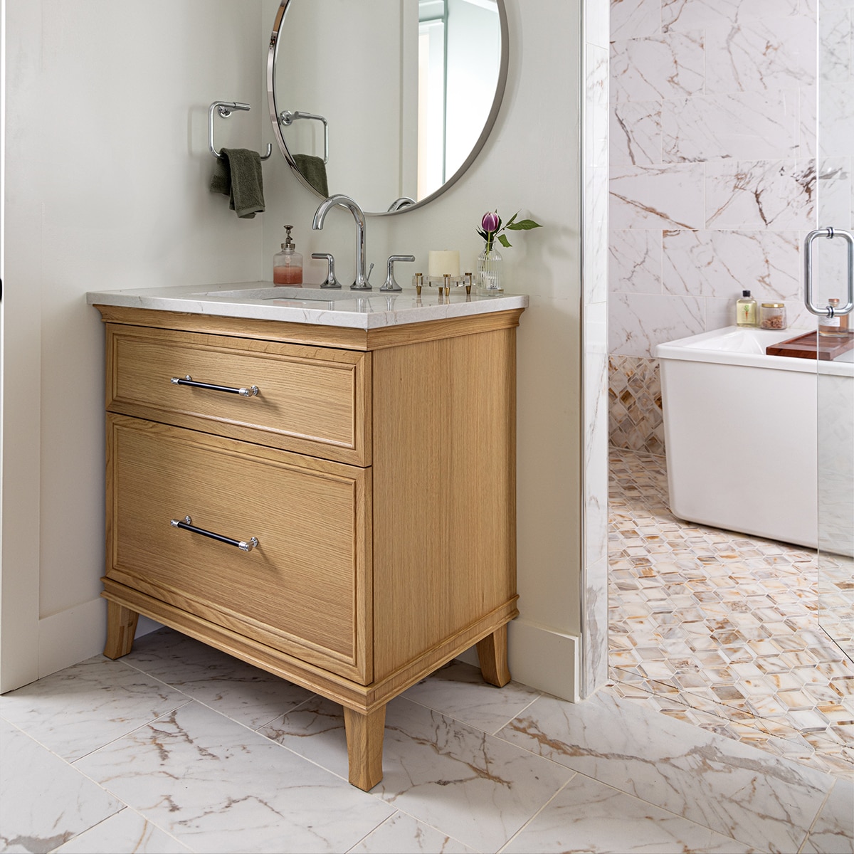 Bathroom with beige quartz on the vanity countertop, white marble look tile on the floor, and in the background a bathtub with white marble look mosaic on the floor and bottom half of the wall and a white marble look tile on the top half of the wall.