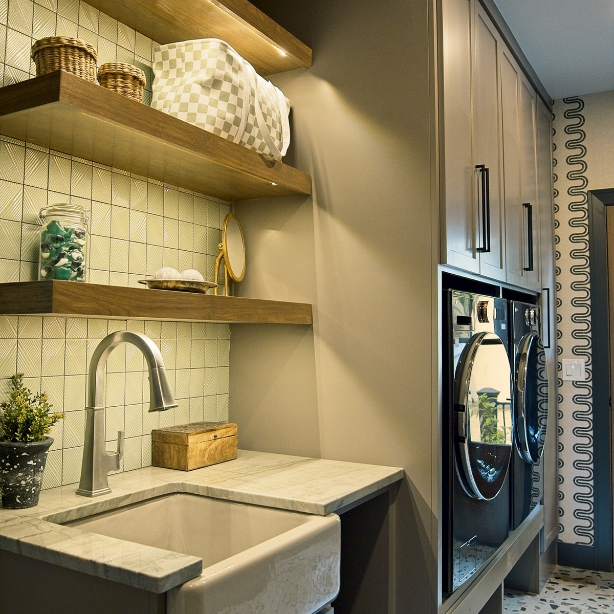 Sideview of mudroom with a green mosaic backsplash, a gray quartzite countertop on the sink, gray cabinets with a washer and dryer., and a white terrazzo look floor tile.
