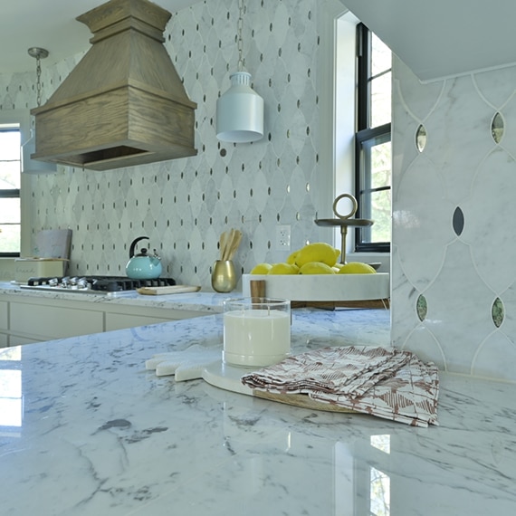 Kitchen with white marble countertops with gray veining, antique mirror & marble mosaic backsplash, wooden hood vent over gas stovetop.