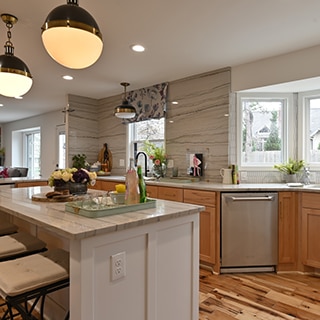 Remodeled kitchen with gray heavily veined quartzite countertop, backsplash, and island, natural wood cabinets, white picket wall tile frames the bay window.