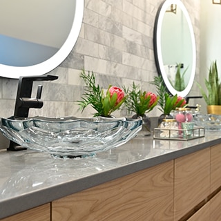 Renovated bathroom vanity with 2 glass vessel sinks, black faucets, gray marble tile backsplash, gray quartz countertop, and wall-mounted round lighted mirrors.