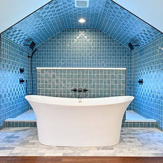 Renovated bathroom with soaker tub on gray marble floor tile, in front of large shower with textured blue geometric tile on the wall and ceiling.