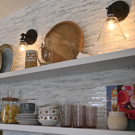 Closeup of coffee nook floating shelves with gray marble mosaic backsplash and wall sconces.