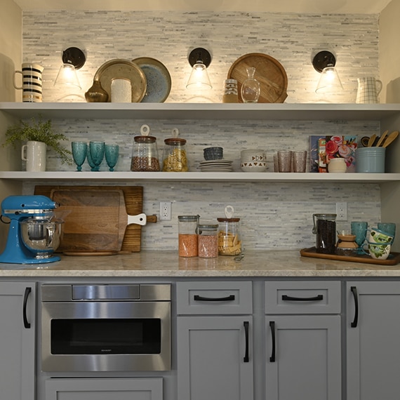 Coffee nook with gray cabinets, tan quartzite countertops, floating shelves, gray marble mosaic backsplash with wall sconces.