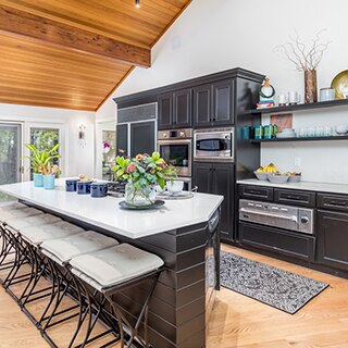 Renovated kitchen featuring white quartz countertop and island, dark wood cabinets, floating shelves, wood slat vaulted ceiling, and large picture windows.