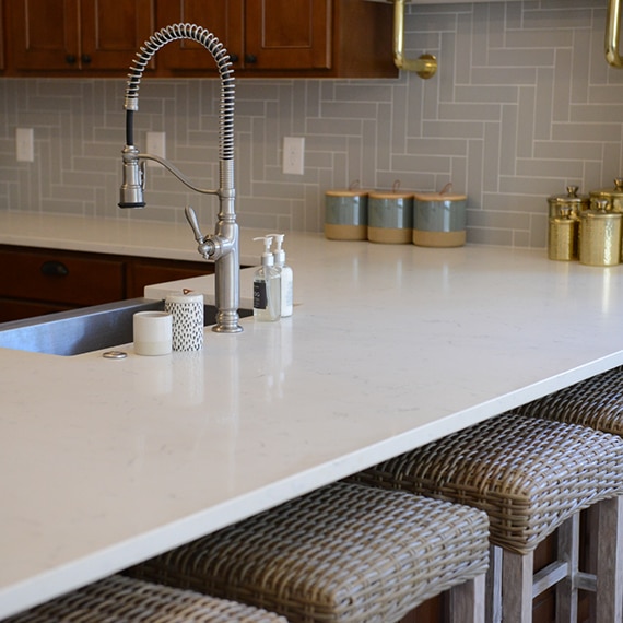 Closeup of kitchen white quartz countertop & peninsula, wicker bar stools, and gray wall tile in a herringbone pattern.