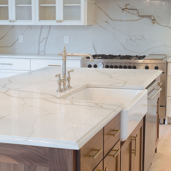 Kitchen with white and light wood cabinetry, marble-look countertops and solid slab marble-look backsplash. Large farmhouse sink in island.