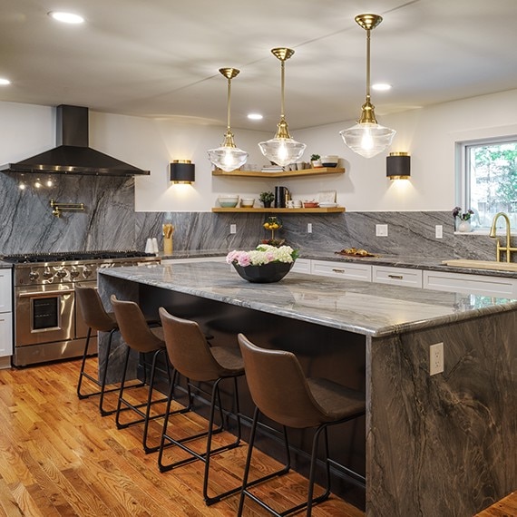 Renovated kitchen featuring gray natural quartzite backsplash, countertops, and waterfall island, floating shelves, white lower cabinets, and brass & glass pendants.