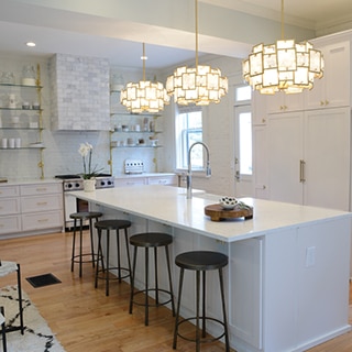 Renovated kitchen with white & gray veining quartz island with built-in sink and pendant lighting, white cabinets, floating shelves, and marble covered vent hood.
