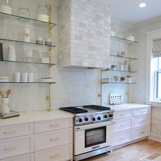 Renovated kitchen with white painted brick wall, stainless steel gas stove/oven, white lower cabinets, floating shelves, and marble covered vent hood.