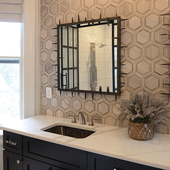 Bathroom vanity with tan & beige limestone hexagon backsplash, white quartz countertop, undermount sink, brushed brass faucet, and black cabinets.