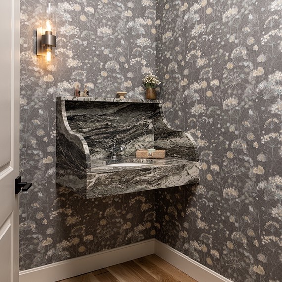 A hall wall vanity with a brown stone marble slab for the sink countertop and backsplash against a gray floral patterned wallpaper.