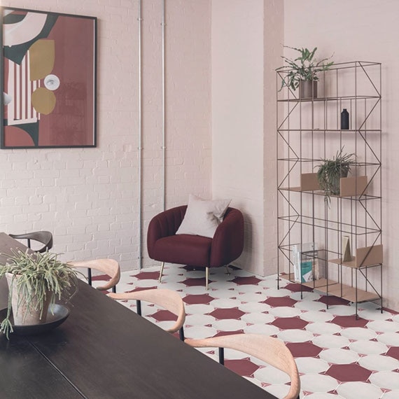 Meeting room with white and maroon hexagon floor tile, dark wood table, light wood chairs, maroon cushioned chair, and open shelving with potted plants.