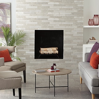 Living room with beige brick fireplace made of textured natural stone marble, gray marble floor tile, gray couch and tan chairs around a wood coffee table.