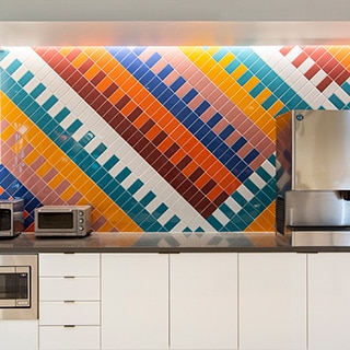Breakroom with wall of bright blue, teal, red, orange, yellow, and white patterned tile, white lower cabinets, and stainless steel kitchen appliances.