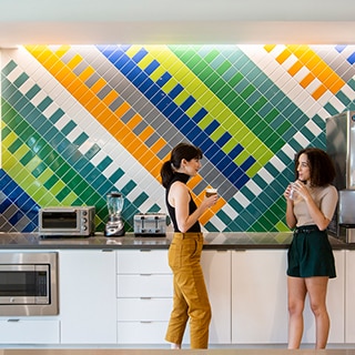 Breakroom with two women conversing in front of wall with bright green, blue, orange, and white patterned tile, white lower cabinets, and stainless steel kitchen appliances.
