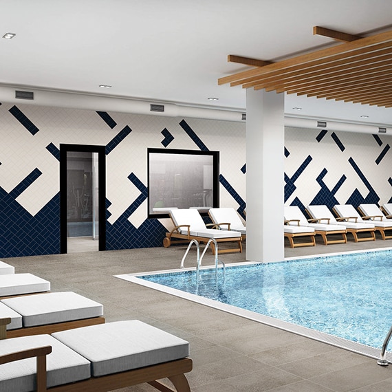 Indoor hotel pool with gray concrete look tile deck, wood lounge chairs with white cushions, and white & navy wall tile in random pattern.