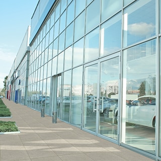 Glass front of a car dealership with sidewalk and walkways of beige porcelain pavers, and showroom with a white and a black car.