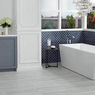 Bathroom with gray floor tile that looks like stone with linear movement, navy blue fan mosaic wall tile, chandelier over soaking bathtub, and gray-blue cabinets.