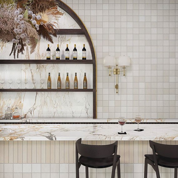 Elegant restaurant bar with porcelain slab countertop that looks like white and golden-brown marble, beige tile wall tile, shelves holding bottles of wine.