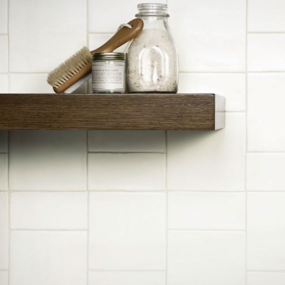 Closeup of bathroom backsplash with intentionally imperfect white wall tile, brush & canister of bath salts on floating woodgrain shelf.