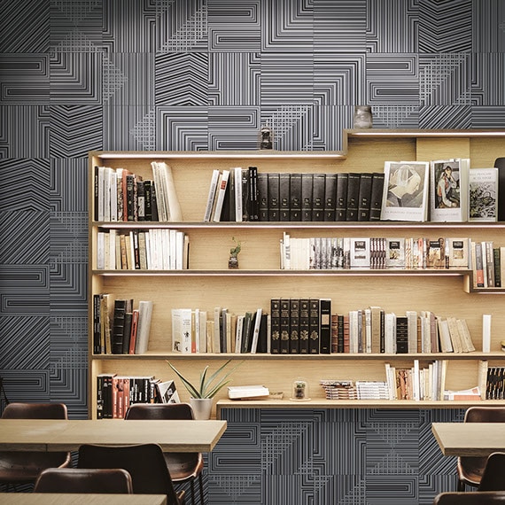 Metallic-look, geometric pattern tile on a library wall, light wood bookshelves with books, tables & chairs.