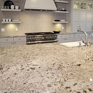 Kitchen with beige & brown granite island with built-in sink, beige limestone backsplash, beige cabinets, hood vent, and floating shelves.