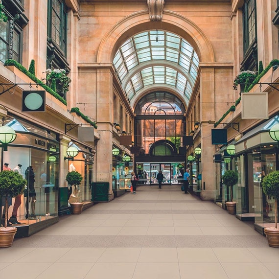 Shopping center with arched skylights, beige & tan floor tile that looks like concrete, glass front boutiques with window dressing and potted plants.
