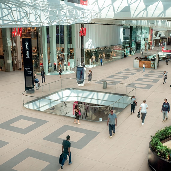 Interior of shopping mall with people walking back & forth, beige floor tile with gray tile accents, and area opened to floor below surrounded by glass railing.