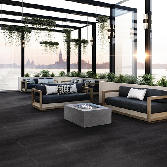 Lakeside restaurant lounge area with black tile flooring, wood frame black sofas around firepits, black metal window & skylight, and view of city skyline.