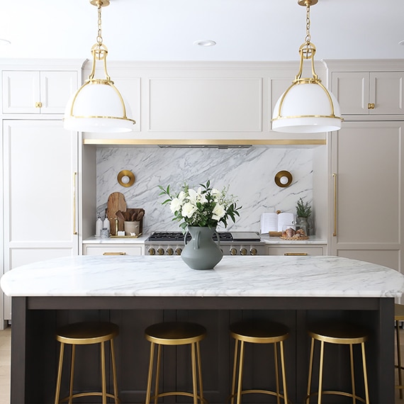 Kitchen with gold accents, white cabinets. center island, sitting stools, and a white marble slab countertop and backsplash.