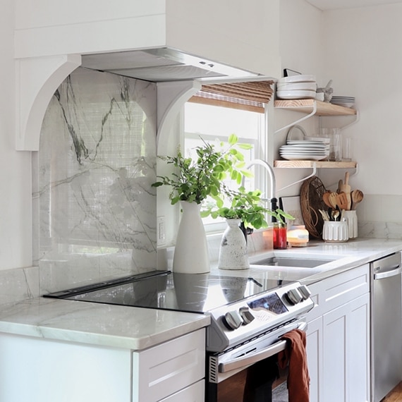 Kitchen with natural quartzite countertops & backsplash, white hood vent, wood floating shelves, and vases with greenery.