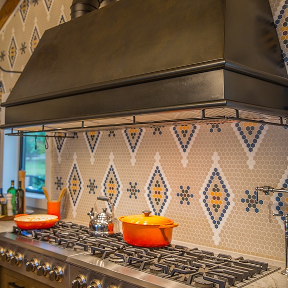 Closeup of kitchen stainless steel gas stovetop & bronze vent hood, light brown quartzite countertop, & island, backsplash of custom tan, white, and tan penny round mosaic tile.