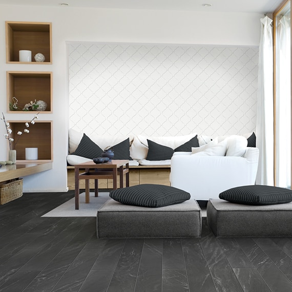Modern living room with white arabesque wall tile, white sofa with gray pillows and natural wood base, black stone look floor tile, and gray ottomans with gray pillows.