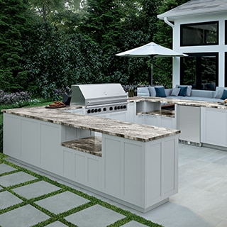 Outdoor kitchen with tan heavily veined quartzite countertops on white cabinets, stainless steel grill, gray stone-look tile flooring, and gray stone-look pavers set in grass.