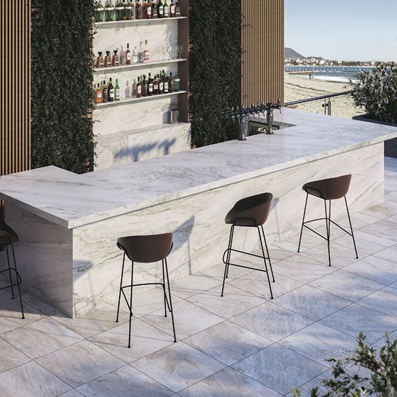 Outdoor hotel bar with gray stone-look floor tile, waterfall island of white quartzite with gray striations, shelves holding bottles of alcohol, beach in the background.