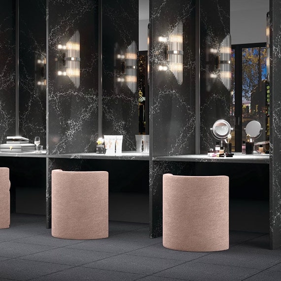 Dressing room with pink vanity stools in front of makeup mirrors divided by black & white quartz walls that look like marble, and dark gray tile flooring.
