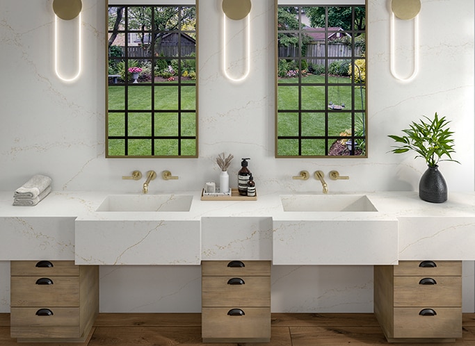 Bathroom with double sinks, white marble look countertops, wood cabinets, and two windows above the sinks showing a backyard.