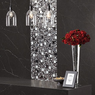 Receiving area with black waterfall quartz slab desk, red flowers in a vase sitting on top. Pendant lighting overhead. Black quartz slab on the walls in the background with vertical stripe of eye-catching glass mosaic.