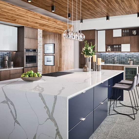 Modern kitchen with marble look, waterfall countertop island, blue glass mosaic tile backsplash, clear pendant lighting, and gray terrazzo floor tile.