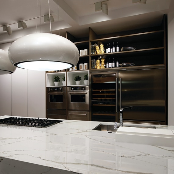 Kitchen island with sink, stove top, and white marble look quart countertop, pendant lighting, stainless steel refrigerator and double oven.